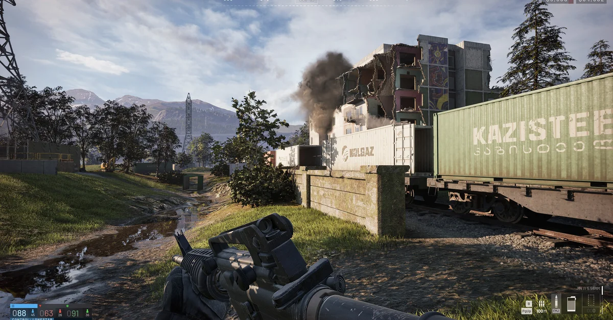 A WARDOGS soldier aims a rifle beside a freight train with a burning apartment block behind